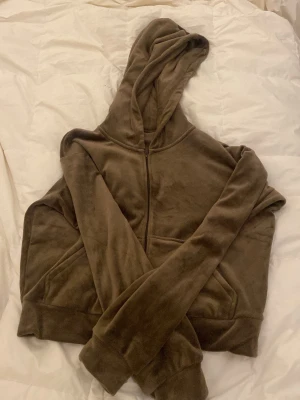 Brun velour hoodie med dragkedja - Mjuk och skön brun hoodie i velourmaterial med dragkedja framtill och stor huva. Hoodien har långa ärmar och ribbade muddar vid ärmslut och nederkant. Perfekt för en chill och avslappnad stil.