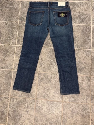 Stone Island jeans - Säljer ett par klassiska blå jeans från Stone Island med rak passform och fem fickor. Jeansen har en snygg tvätt och det ikoniska Stone Island-märket på bakfickan. Tillverkade i slitstarkt denim med kontrastsömmar för en cool look.