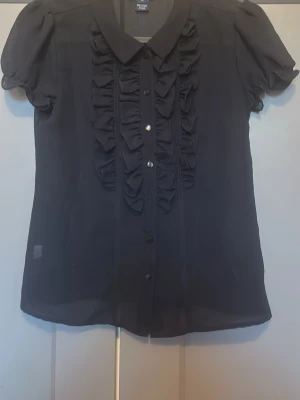 Marinblå blus med volang från Ralph Lauren - Stilren Marinblå blus från Ralph Lauren med korta puffärmar och dekorativa volanger framtill. Blusen har klassisk krage och knäppning hela vägen ner. Tunt, lätt material som ger en luftig känsla och snygg passform.