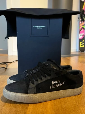 Svarta Saint Laurent sneakers - Svarta låga sneakers från Saint Laurent med vit logga på sidan och svart textil samt läderdetaljer. Skorna har svarta skosnören och en vit, något smutsad sula. Klassisk och stilren design med Saint Laurent Paris-detaljer. Box och dustbag ingår! 