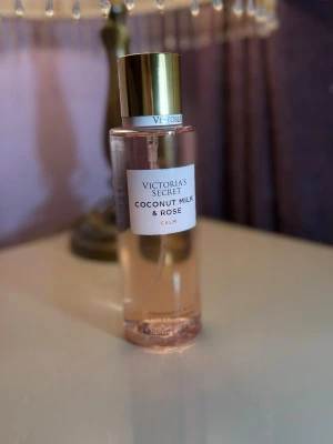 Victoria's Secret Coconut Milk & Rose - Fräsch parfym från Victoria's Secret med doft av kokosmjölk och ros. Flaskan är cylinderformad i transparent plast med ljusrosa vätska och har ett elegant guldfärgat lock. Perfekt för dig som gillar blommiga och exotiska dofter.