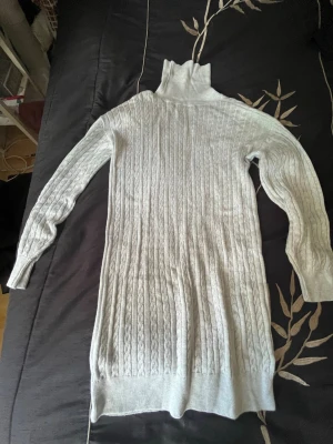 Ljusgrå poloklänning kabelstickad - Säljer en  kabelstickad poloklänning i ljusgrått. Klänningen har hög polokrage, långa ärmar och ribbade muddar. Perfekt för lager på lager och riktigt mysig under kalla dagar. Materialet känns mjukt och stretchigt. Inte använt så mycket å nu är de för liten.