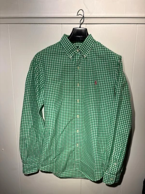 Grön rutig skjorta från Ralph Lauren - Snygg grön och vit rutig skjorta från Ralph Lauren med klassisk button-down krage. Tillverkad i mjuk bomull, perfekt för en clean och fräsch look. Skjortan kommer passa perfekt nu under sommaren och de soliga dagarn, men även under höst och vinter när det blir lite mörkt, kommer den lysa upp och skina. Passar perfekt till både kavaj men även att hänga en tröja över axlarna. Skjortan är i fläckfritt skick och har inga deffekter. 