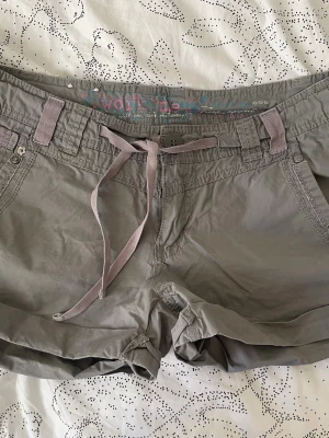 Lågmidjade grå shorts med snörning  - Snygga grå shorts med snörning i midjan och praktiska fickor både fram och bak. Shortsen har bälteshällor och dekorativa knappar på bakfickorna. Perfekta för varma dagar och en avslappnad stil. Står ingen storlek men skulle säga S
