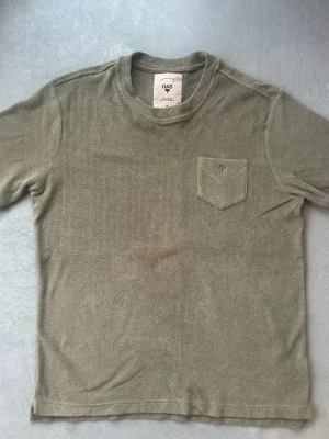 Olivgrön frotté t-shirt från OAS - Säljer en olivgrön t-shirt från OAS i mjuk frotté. T-shirten har korta ärmar, rund halsringning och en bröstficka med diskret broderad detalj. Perfekt för chill dagar eller efter ett dopp. Enkel och stilren design. Aldrig använd