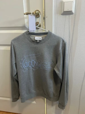 Grå sweatshirt från Lacoste - Snygg grå sweatshirt från Lacoste med stort broderat krokodillogo och text på bröstet. Klassisk rund halsringning och ribbade muddar vid ärmslut och nederkant. Perfekt för en avslappnad och trendig look.