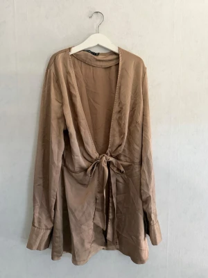 Beige omlottopp med knytband - Snygg beige omlottopp i satinliknande material med långa ärmar och djup v-ringning. Toppen har ett knytband i midjan som ger en feminin siluett. 