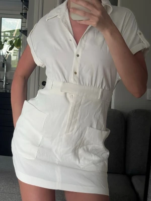 Vit kort skjortklänning med fickor - Snygg vit kort skjortklänning med krage, knappar framtill och stora fickor på sidorna. Klänningen har korta ärmar med knappdetalj och markerad midja. Perfekt för en fräsch och avslappnad look.