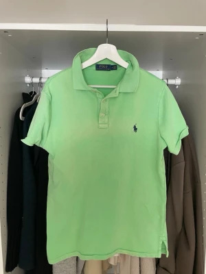 Grön pikétröja från Polo Ralph Lauren - Snygg ljusgrön pikétröja från Polo Ralph Lauren med klassisk krage och knappar framtill. Tröjan har korta ärmar och den ikoniska broderade loggan på bröstet. Perfekt till jeans eller shorts för en fräsch look.