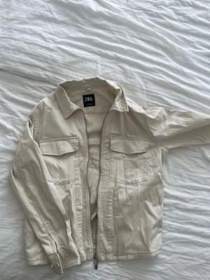 Beige overshirtjacka från Zara - Stilren beige overshirt jacka från Zara med två bröstfickor och dragkedja framtill. Overshirten har klassisk krage och raka ärmar med knapp vid ärmslut.  