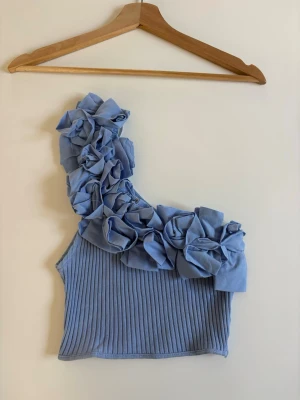 Blå oneshoulder topp med volanger från Zara - Snygg ljusblå oneshoulder topp från Zara med stora volanger längs axeln. Toppen är ribbad och har en croppad passform som ger en trendig look. Perfekt för dig som vill sticka ut med en unik detalj på din outfit.