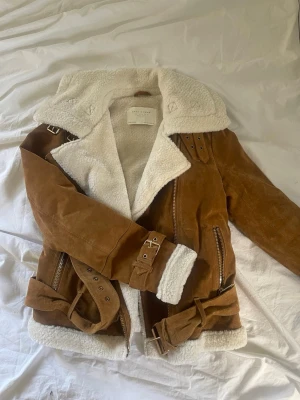 Brun shearlingkappa från Zara - Säljer en brun shearlingkappa från Zara i mocka med vit fuskpälsfoder. Kappan har bred krage, dragkedjor, bältesdetaljer vid ärmslut och midja samt stora fickor. Perfekt för kalla dagar och riktigt snygg till jeans eller kjol.
