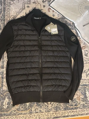 Canada goose cardigan black label - Snygg svart pufferjacka från Canada Goose med quiltad front och stickade ärmar samt rygg. Jackan har dragkedja framtill, ribbade muddar och en patch med logga på ärmen. Perfekt för kalla vinterdagar och riktigt stilren look. Påsar och tags tillkommer!