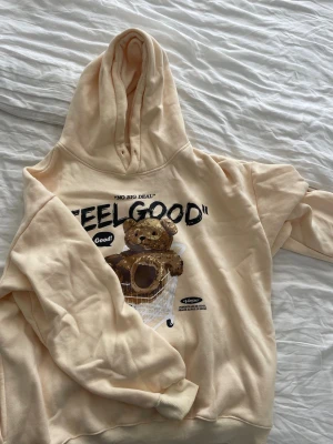 Beige hoodie med björntryck - Säljer en beige hoodie med stor huva och tryck av en nallebjörn i en kundvagn samt texten 'FEELGOOD' på bröstet. Hoodien har avslappnad passform och är gjord i mjukt bomullsmaterial. 