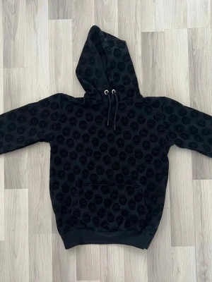 Svart hoodie med mönster - Snygg svart hoodie med ett all-over mönster. Hoodien har huva med dragsko och en stor magficka framtill. Tillverkad i mjukt material som känns skönt mot huden. Perfekt för dig som vill ha en exklusiv streetwear-look.