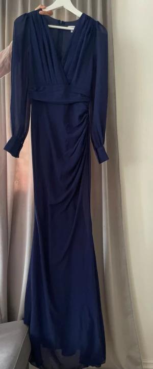 Marinblå långklänning - Elegant marinblå långklänning med långa luftiga ärmar. Klänningen har markerad midja och draperad detalj på sidan som ger en snygg siluett. Tillverkad i ett mjukt, lätt tyg som faller fint.