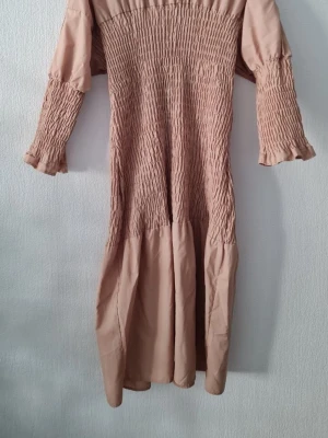 Beige långklänning med smock - Trendig beige långklänning med smockdetaljer över hela överdelen och ärmarna. Klänningen har långa ärmar med volangkant och en flowy nederdel. Perfekt för dig som gillar en avslappnad men ändå chic stil. Extra knapp ingår. Storlek 42, men passar även mindre och större storlekar. 🤎 100% bomull. Nypris: 599 kr. Saknar prislapp men den är hrlt ny och har aldrig kommit till användning. 