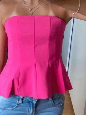 Rosa axelbandslös korsettopp - Snygg rosa korsettopp med axelbandslös design och figurnära passform. Toppen har en lätt utsvängd nederkant och markerade sömmar framtill för en trendig look. Perfekt att styla med jeans eller kjol.