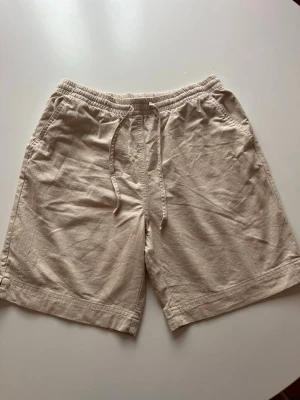 Beige linneshorts med dragsko - Säljer ett par ljusbeige shorts i linne med avslappnad passform och elastisk midja med dragsko. Perfect till sommaren.Shortsen har två sidofickor och är perfekta för varma dagar. Materialet är luftigt och ger en chill vibe.de har aldrig använts. 