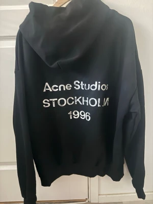 Svart hoodie Acne Studios Stockholm - Svart hoodie från Acne Studios med stor vit text på ryggen där det står 'Acne Studios STOCKHOLM 1996'. Klassisk passform med huva och långa ärmar. Perfekt för dig som gillar streetwear och vill ha en stilren men ändå statement look.