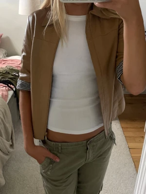 Beige skinnjacka från MGN - Stilren beige skinnjacka  med trekvartsärmar och klassisk krage. Jackan har rutiga svarta och vita detaljer på ärmsluten med knapp, samt en mjuk, glansig innerfoder. Perfekt för dig som vill ha en unik och trendig look.