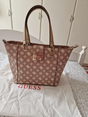 Beige monogram handväska från Guess - Snygg beige handväska från Guess med klassiskt monogrammönster i vitt. Väskan har två långa handtag, guldfärgade metalldetaljer och en dekorativ logotyp framtill. Tillverkad i konstläder och har en praktisk dragkedjeficka på baksidan.