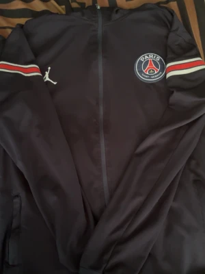 Svart PSG träningsjacka från Jordan - Snygg svart träningsjacka från Jordan x PSG med dragkedja, vita och röda ränder på ärmarna samt PSG-logga på bröstet. Jackan har även Jordan-loggan på ena sidan. Perfekt för dig som gillar fotboll och streetwear.