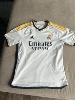 Real Madrid matchtröja Adidas vit - Säljer en vit Real Madrid matchtröja från Adidas med gula och mörkblå detaljer. Tröjan har klubbmärke och Adidas-logga på bröstet samt 'Emirates Fly Better' tryckt framtill. Tillverkad i lätt och ventilerande material, perfekt för fotbollsträning eller match.