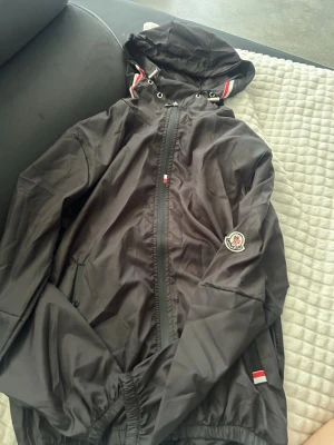 Moncler windbraker - Snygg svart vindjacka från Moncler med huva och dragkedja framtill. Jackan har Moncler-logga på ärmen och detaljer i rött, vitt och blått på huvan och fickan. Tillverkad i lätt, glansigt material som står emot vind.