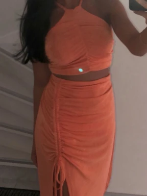 Orange croptop med rynkade detaljer - Säljer en snygg orange croptop med halterneck och rynkade detaljer framtill på kjolen . Toppen är figurnära och har en kort passform som visar magen. Kjolen är väldigt strechtig också. Perfekt för dig som gillar trendiga och färgstarka plagg.