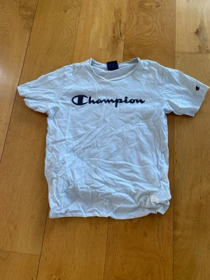 Vit Champion t-shirt med logga - Vit t-shirt från Champion med klassisk svart logga tryckt över bröstet och liten logga på ärmen. T-shirten har rund halsringning och korta ärmar. Tillverkad i mjuk bomull för en skön och avslappnad stil.