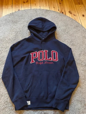 Mörkblå hoodie från Polo Ralph Lauren - Snygg mörkblå hoodie från Polo Ralph Lauren med stor röd och vit POLO-logga broderad på bröstet. Klassisk känguruficka och huva med dragsko. Perfekt för en avslappnad och sportig stil.