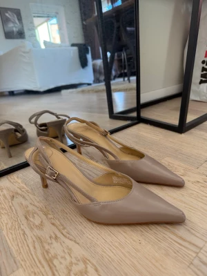 Beige slingback pumps från H&M - Stilrena klackar i beige från H&M med spetsig tå och låg klack. Skorna har en öppen häl med justerbart spänne och är tillverkade i ett slätt, syntetiskt material. 