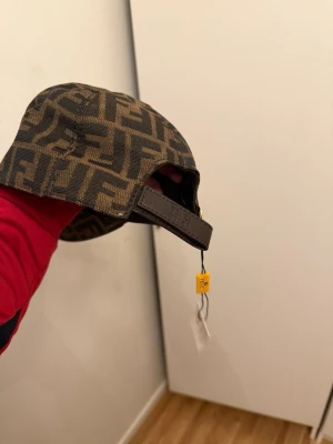 Fendi keps med logomönster - Säljer en snygg keps från Fendi med klassiskt brunt och svart logomönster över hela kepsen. Justerbar rem baktill i läder och cool, böjd skärm. Perfekt för dig som vill sticka ut med exklusiv stil och streetwear-vibe.