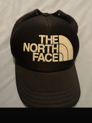 Svart keps från The North Face - Snygg svart keps från The North Face med stort vitt logotryck framtill. Klassisk trucker-modell med mesh på sidorna och justerbar snapback baktill. Perfekt för en avslappnad streetwear-look.