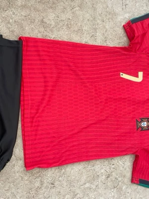 Portugal matchtröja röd nummer 7 - Säljer en röd Portugal matchtröja från Nike med guldigt nummer 7 på både fram- och baksidan. Tröjan har korta ärmar, gröna och svarta detaljer vid axlarna och är tillverkad i ett lätt, ventilerande funktionsmaterial. Perfekt för fotbollsträning eller att bära till vardags.