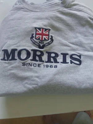 Grå sweatshirt från Morris XS - Cool grå sweatshirt från Morris med broderad logga och Union Jack på bröstet. Tröjan har rund hals och ribbade muddar. Perfekt för dig som gillar klassisk stil med en twist. Materialet är mjukt och bekvämt, idealiskt för chill dagar.