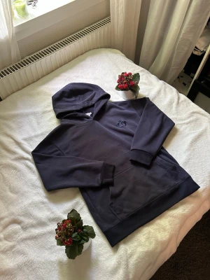 Mörkblå CD hoodie - Snygg mörkblå hoodie med huva och känguruficka framtill. Broderad logga i samma färg på bröstet ger en clean look. Tillverkad i mjukt bomullsmaterial som är skönt mot huden. Perfekt för chill dagar eller när du vill ha en enkel men stilren outfit.