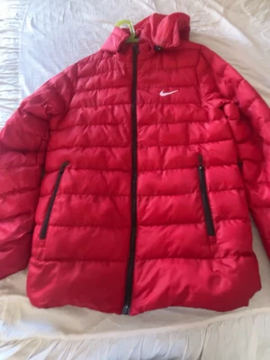 Röd pufferjacka från Nike - Snygg röd pufferjacka från Nike med svart dragkedja och två stora fickor framtill. Jackan har huva och den klassiska Nike-loggan på bröstet. Perfekt för kalla dagar och ger en sportig look. Tillverkad i vadderat material som håller värmen.