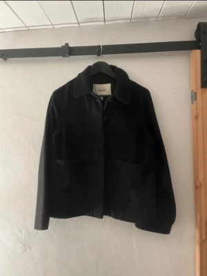 Svart kappa från AWARE - Stilren svart jacka från AWARE i mjuk material . Klassisk krage, stora framfickor och knäppning framtill. Perfekt för dig som gillar en enkel men trendig look. Passar till det mesta och är lätt att matcha.