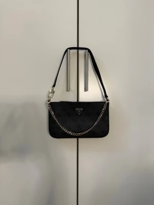 Svart handväska från Guess - Snygg svart handväska från Guess med diskret logomönster och silverfärgad kedjedetalj framtill. Väskan har en avtagbar axelrem och en praktisk ytterficka med dragkedja på baksidan. Perfekt storlek för det viktigaste och stilren design.
