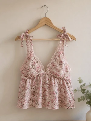 Blommig spetstopp med rosetter - Söt topp i ljusrosa spets med blommigt mönster och tunna axelband som knyts med rosetter. Djup urringning med spetskant och smock i ryggen för skön passform. Materialet är genombruten spets med romantisk känsla.