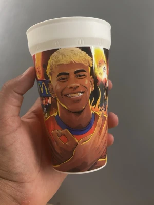 McDonald's samlarmugg med fotbollsmotiv - Cool samlarmugg från McDonald's med färgglad tecknad design av en fotbollsspelare i röd och blå tröja. Muggen handlar om lamin yamal. Muggen har texten 'Lamine Yamal' i eldiga bokstäver och är gjord i plast med vit kant upptill. Perfekt för dig som gillar fotboll och unika samlarprylar.