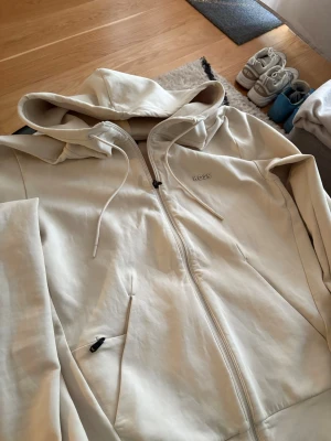 BOSS saggy hoodie  - Snygg beige hoodie från BOSS med dragkedja framtill och två fickor. Klassisk huva med dragsko och diskret BOSS-logga på bröstet. Tillverkad i mjukt material som känns skönt mot huden. Perfekt för en avslappnad och stilren look.