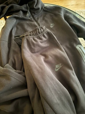 Svart Nike tech fleece  - Mysigt svart loungewear set från Nike med matchande hoodie och byxor. Hoodien har dragkedja och byxorna har resår i midjan med snörning. Båda plaggen har Nike-logga framtill. Perfekt för chill dagar hemma eller när du vill vara bekväm.