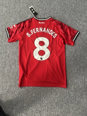 Manchester United matchtröja Adidas - Säljer en röd Manchester United matchtröja från Adidas med B. Fernandes och nummer 8 på ryggen. Tröjan har vita och svarta detaljer, klubbmärke på bröstet och Snapdragon som sponsor. Tillverkad i lätt och ventilerande material, perfekt för fotboll eller träning.