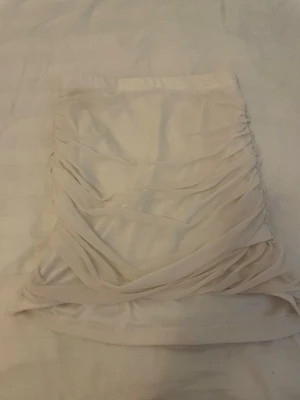 Vit kortkjol med rynkade detaljer - Säljer en vit kortkjol med snygga rynkade detaljer på sidorna. Kjolen har en figurnära passform och är tillverkad i ett mjukt och stretchigt material som känns bekvämt. Perfekt för dig som gillar en stilren och trendig look.