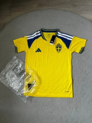 Sverige matchtröja Adidas gul/blå - Säljer en officiell Sverige matchtröja från Adidas i gult med blå detaljer och vita ränder på axlarna. Tröjan har Sveriges landslagsemblem på bröstet och klassisk V-ringning. Tillverkad i lätt och ventilerande funktionsmaterial.Den är i bästa kvalité och helt ny, därför är den 10 av 10 i skick.Väldigt fin och bra för sommaren och för träningen. Fraktar så fort du har köpt🚚📦Aktiv 24/7✅ Hör av dig via frågor⁉️