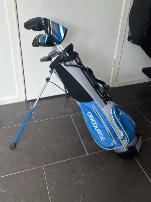 Blå golfset från OnCourse - Säljer ett komplett golfset från OnCourse med blå och grå bärbag, matchande headcovers och fem klubbor. Setet innehåller driver, en järn 9, en sandwedge och en putter. Perfekt för dig som vill börja spela golf eller behöver ett extra set. Snygg design och praktisk bärrem.
