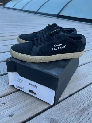 Svarta Saint Laurent sneakers - Svarta sneakers från Saint Laurent med vit broderad logga på sidan. Skorna har svart ovandel i mocka och läder, svarta skosnören och en beige sula i gummi. Klassisk låg modell med stilren design och exklusiv känsla.